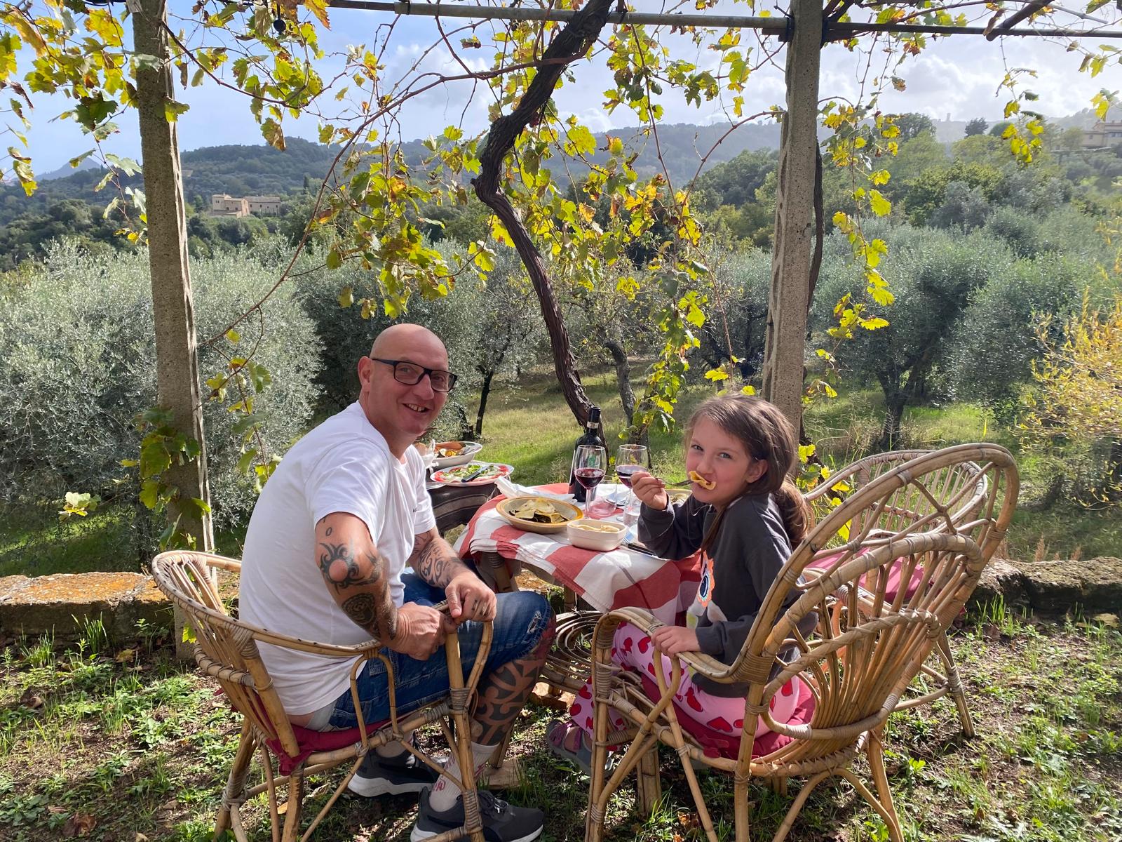 Andrea und seine Tochter beim Mittagessen an einem Tisch im Freien, Olivenbäume und Hügel im Hintergrund