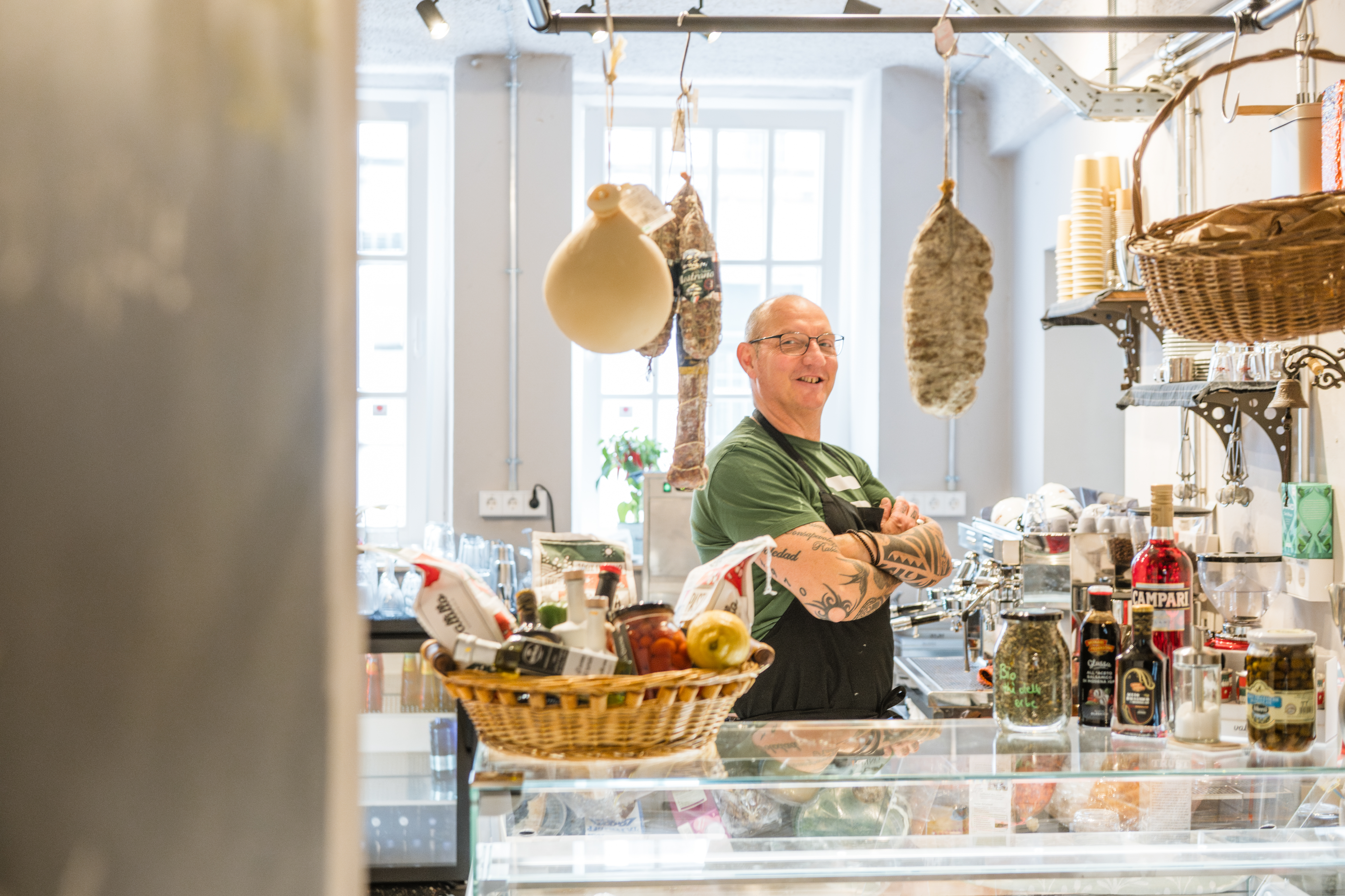 Andrea hinter der Feinkosttheke im Zuckerspeicher — lächelnd zwischen hängendem Salami und Antipasti