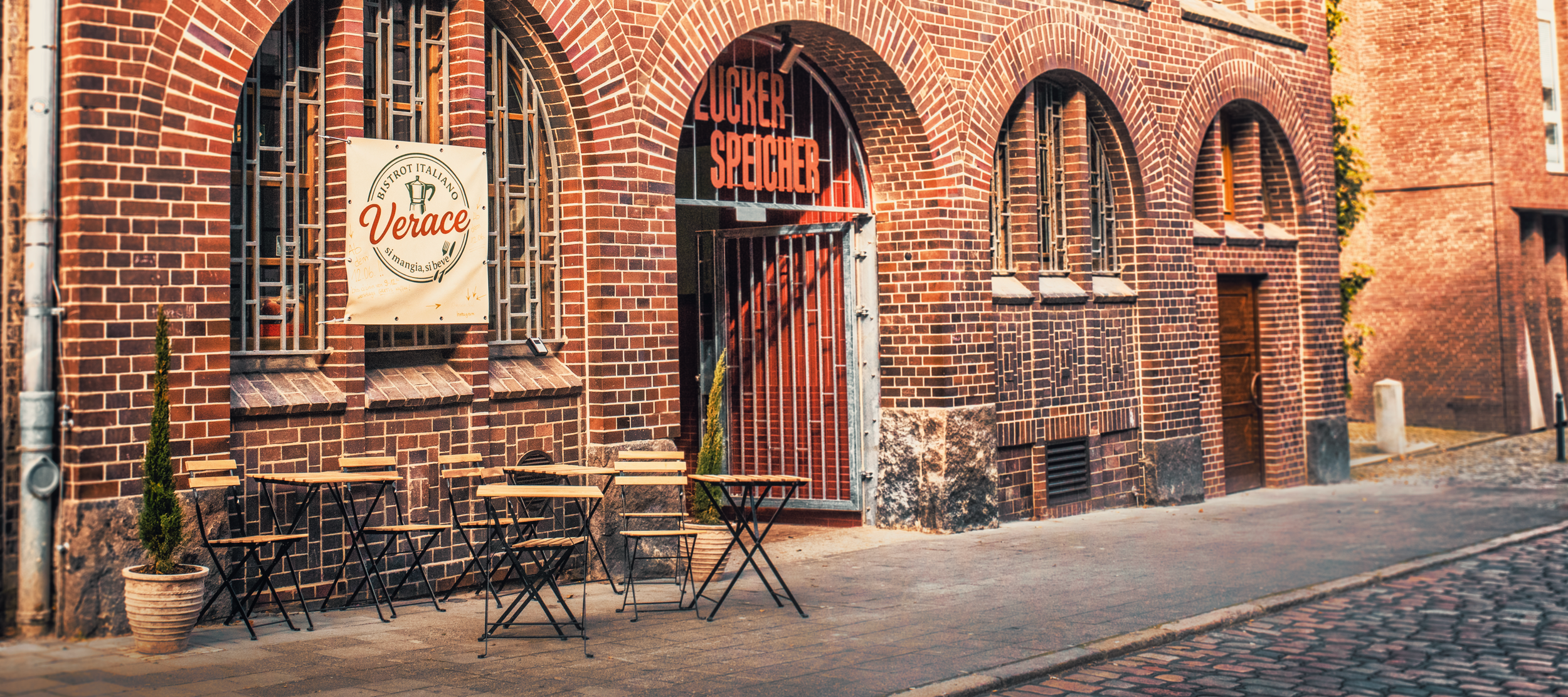 Verace im Zuckerspeicher — Gäste an Bistrotischen vor dem Backsteinbau im Abendlicht